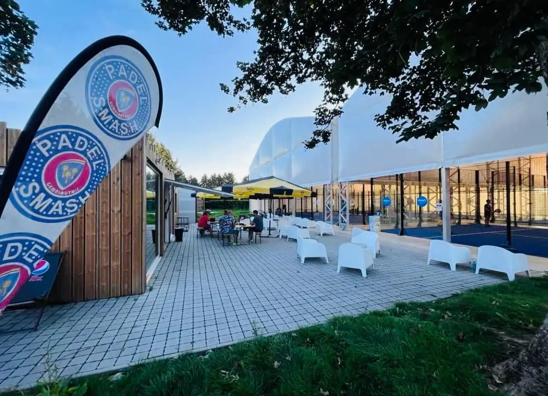 La terrasse du Padel Smash Gembloutois en fin de journée : l'oriflamme du club, les tables et fauteuils, et les terrains de padel couverts sous leur structure blanche.