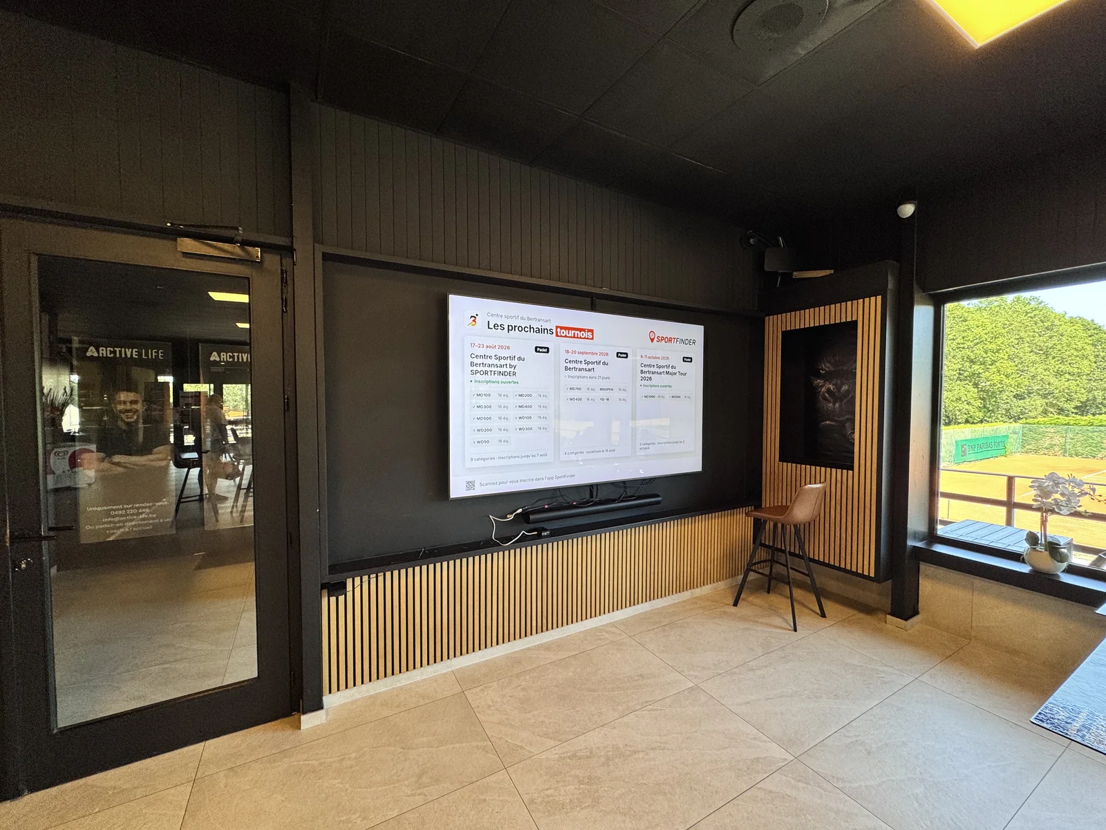 Salon d'un club de padel avec un écran TV SportFinder affichant les prochains tournois, les terrains visibles par la baie vitrée