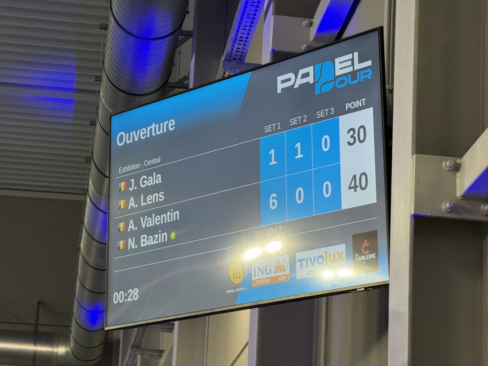 Écran suspendu au-dessus d'un terrain de padel affichant le score du match en cours : sets, points et chronomètre
