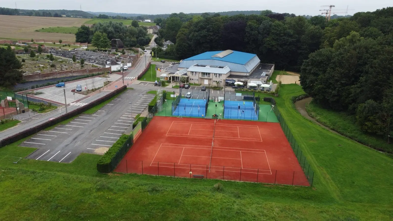 Vue aérienne d'un centre multisport : courts de tennis en terre battue, pistes de padel occupées par des joueurs, terrain de basket et hall couvert.