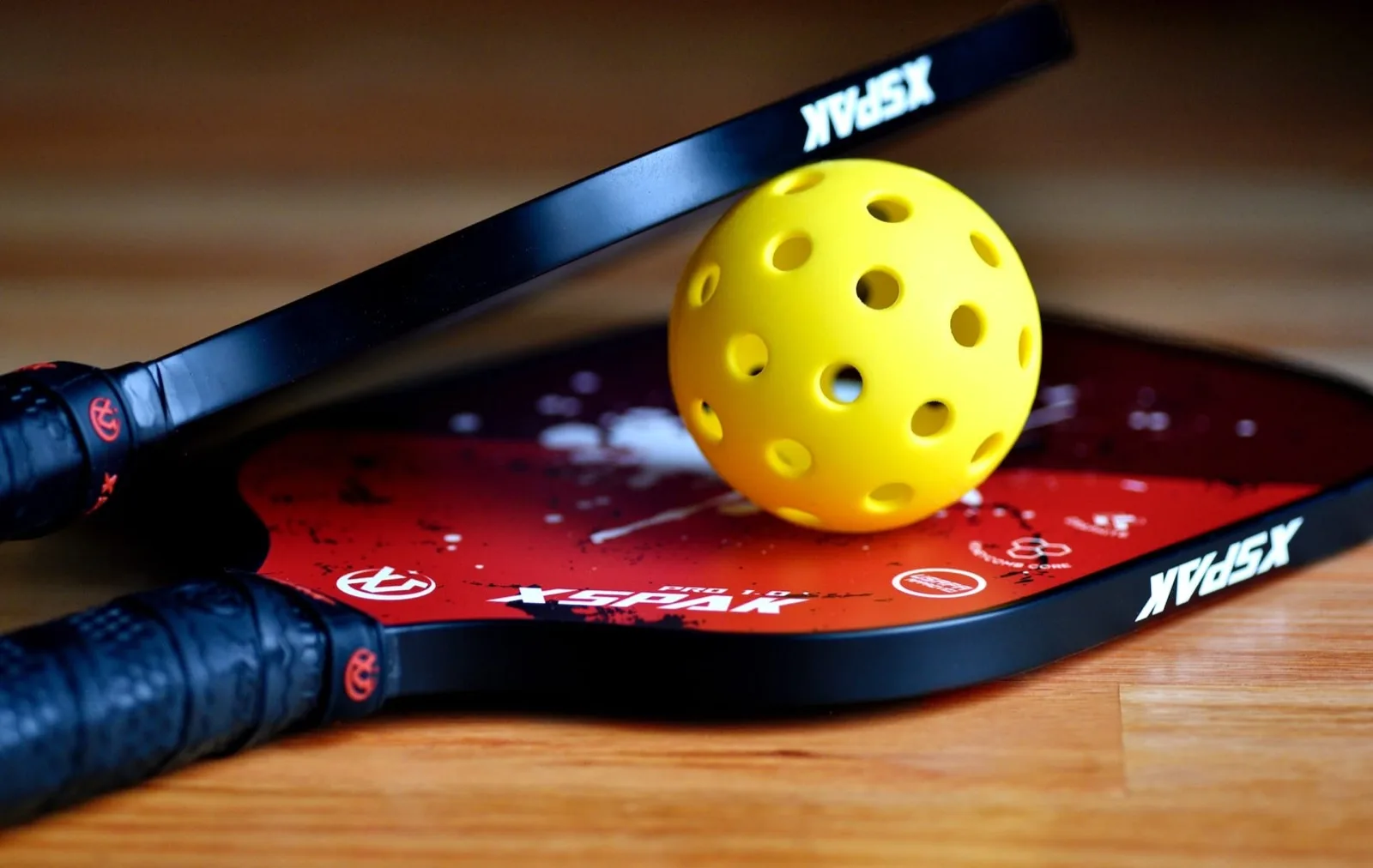 Deux raquettes de pickleball et une balle jaune perforée, posées sur un parquet.