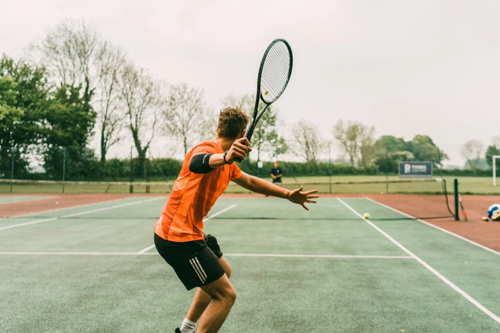 Un joueur de tennis frappe un coup droit, raquette au-dessus de la tête, sur un court extérieur.